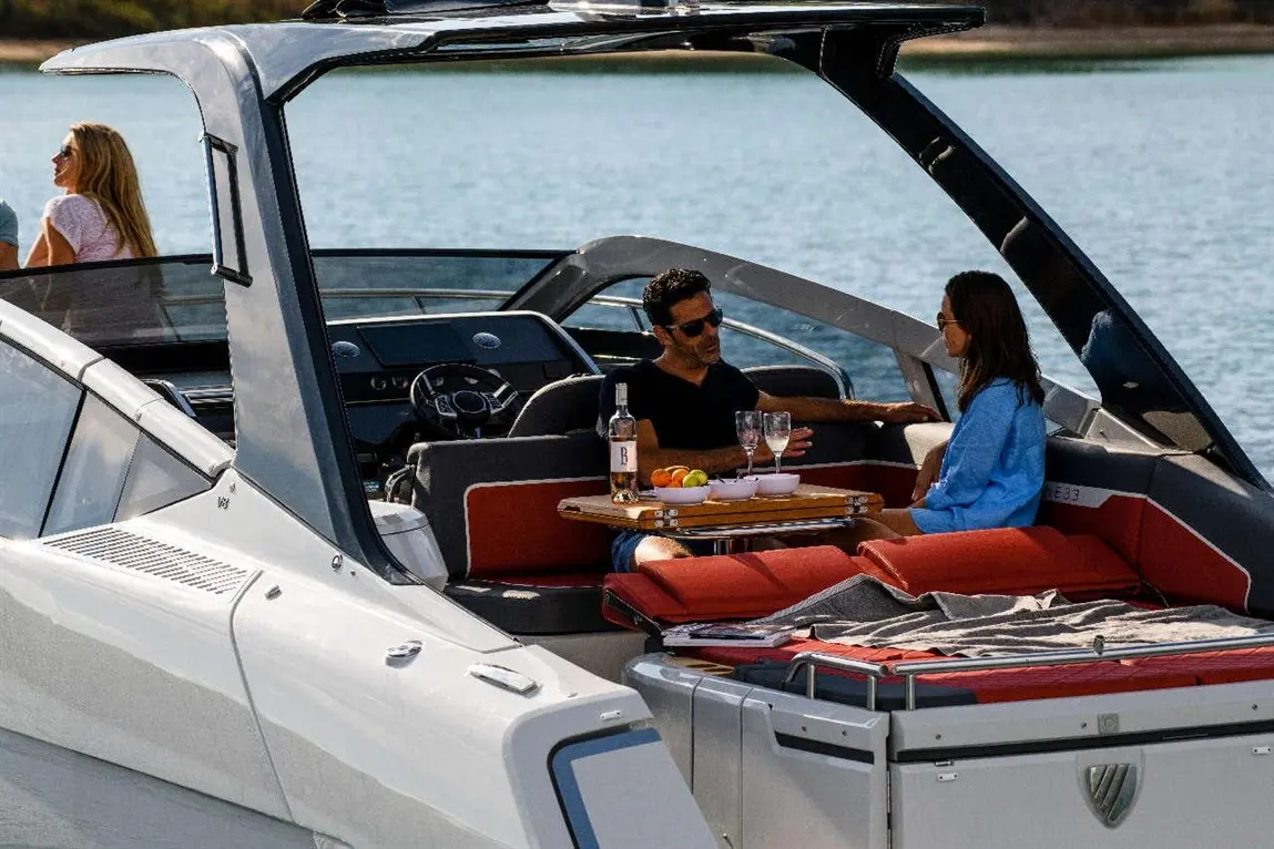 Couple enjoying drinks on a sleek boat, with a table of fruit and glasses, overlooking calm water in a sunny setting.
